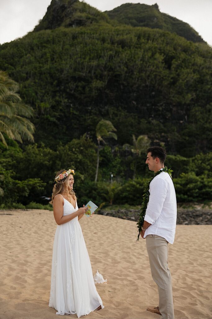 A bride and groom smile at one another while standing on a beach in Hawaii with a large green mountain behind them, the bride is holding a letter from the groom to read