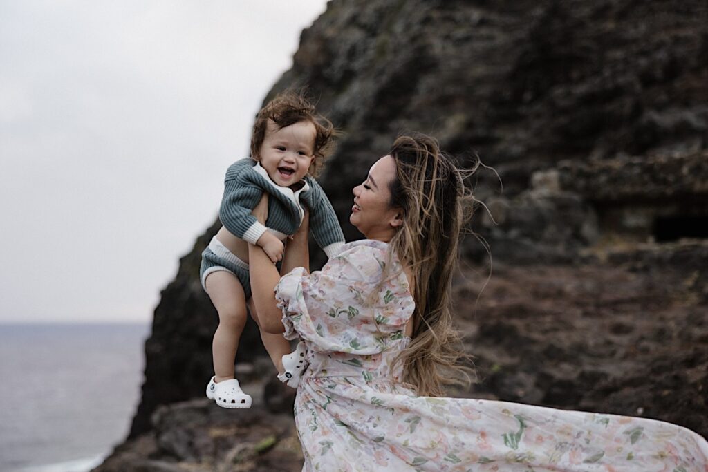 A child laughs while being held up by their mother during a family session at Makapuu Lookout on Oahu, behind them is a large rock formation and the ocean