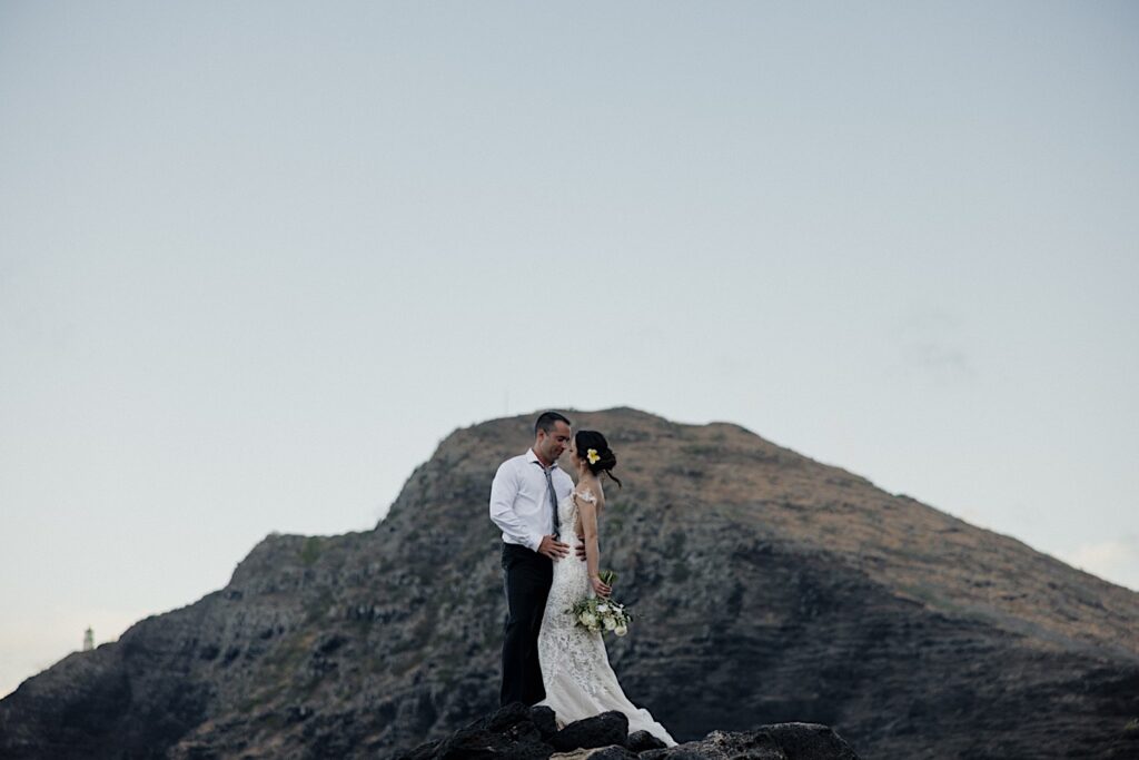 A bride and groom stand together and embrace while on a cliff, behind them is a mountain