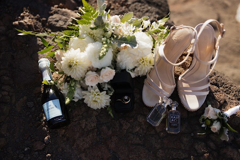 Detail photo of a bridal bouquet, shoes, wedding rings, perfume, and champagne all laying on a rock