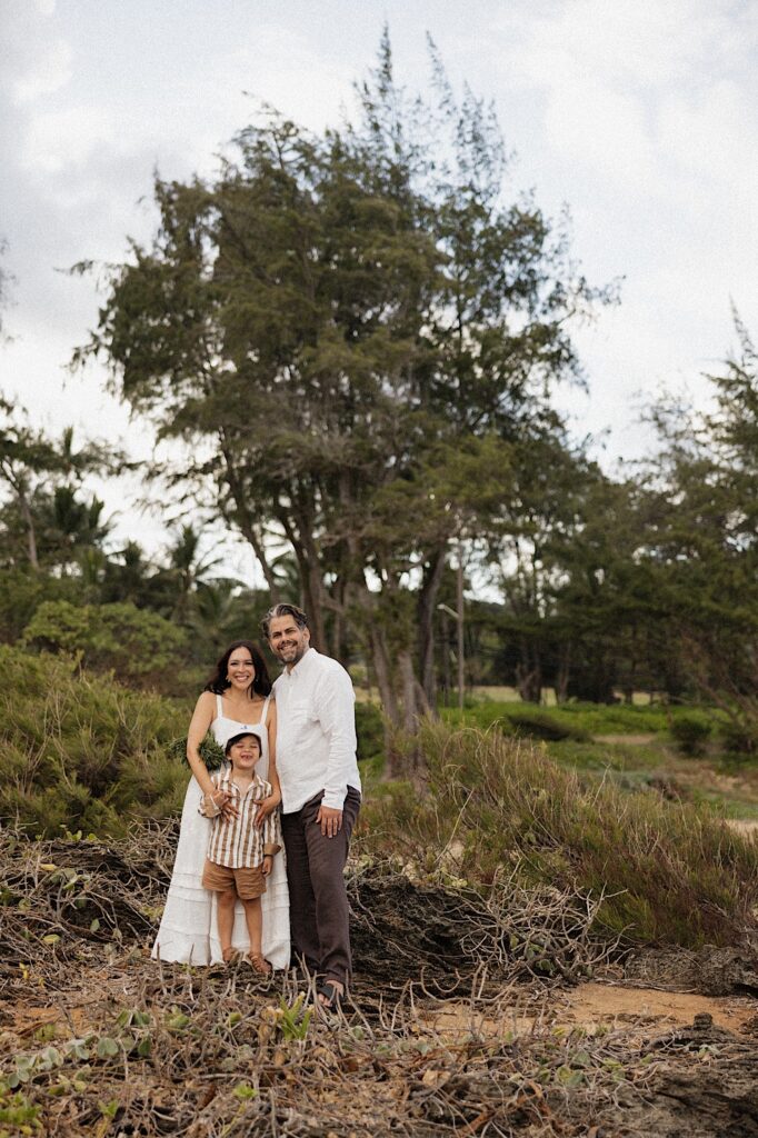 A family of 3 stand together and smile at the camera while standing on a hill in front of a forest in Hawaii