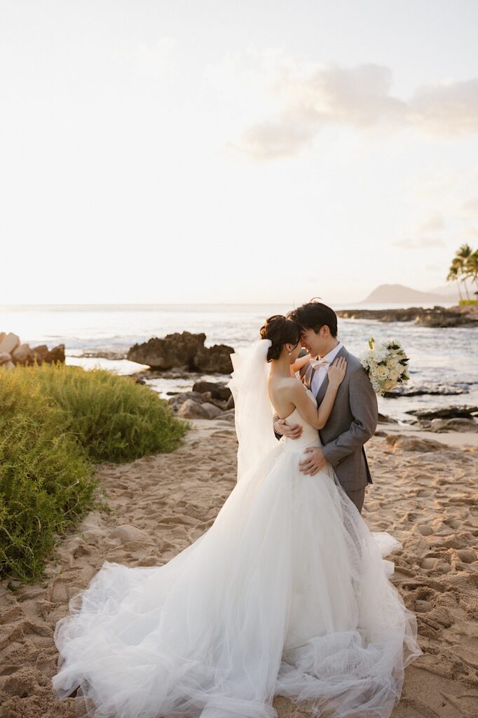 A bride and groom on a beach in Hawaii embrace one another and touch their foreheads together with the ocean and sunset behind them