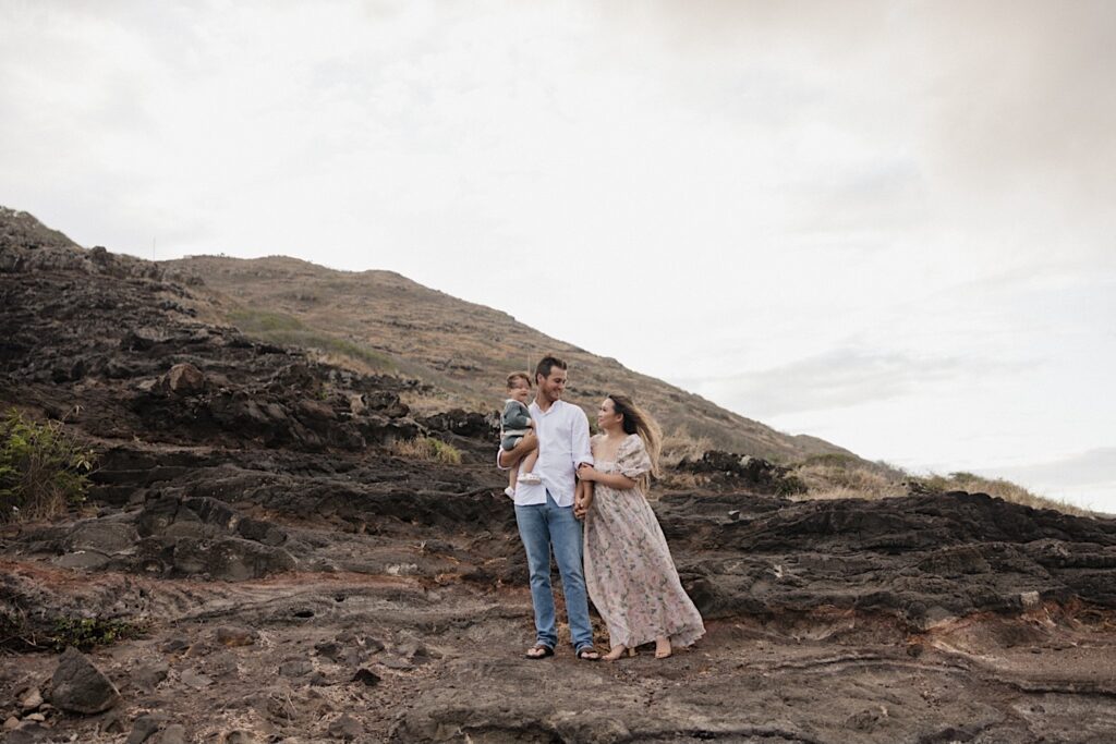 A mother and father stand side by side holding hands atop Makapuu Lookout on Oahu during their family session, the father has their young child resting in one of his arms