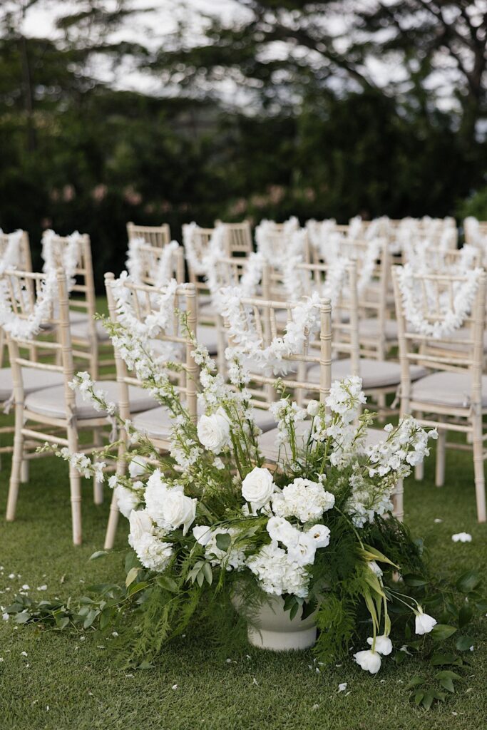 White flowers in a stone vase sit on the ground next to white chairs with white flower leis on them for a wedding ceremony