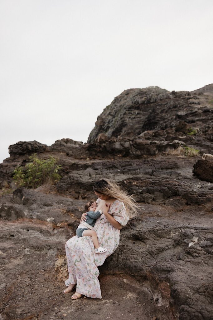 A young child breastfeeds from their mother while she sits on a large rock formation