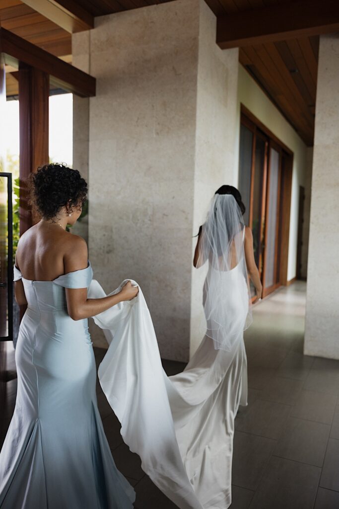 A bride walks away from the camera as a bridesmaid carries the tail end of her dress for her
