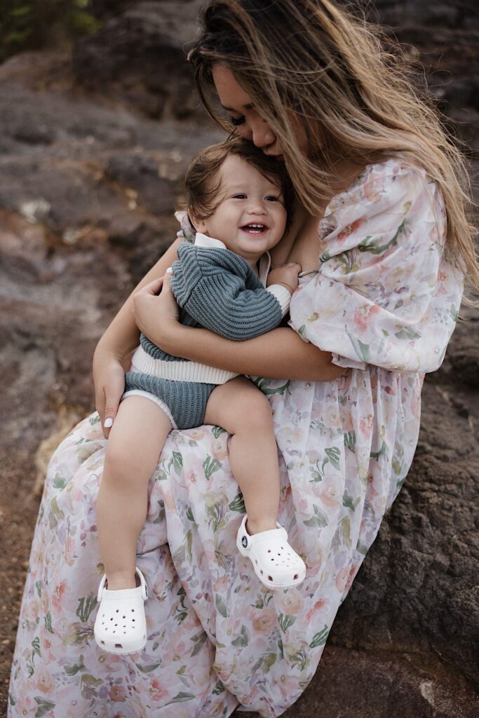 A child smiles at the camera while being held and cuddled by their mother who is sitting on a rock formation