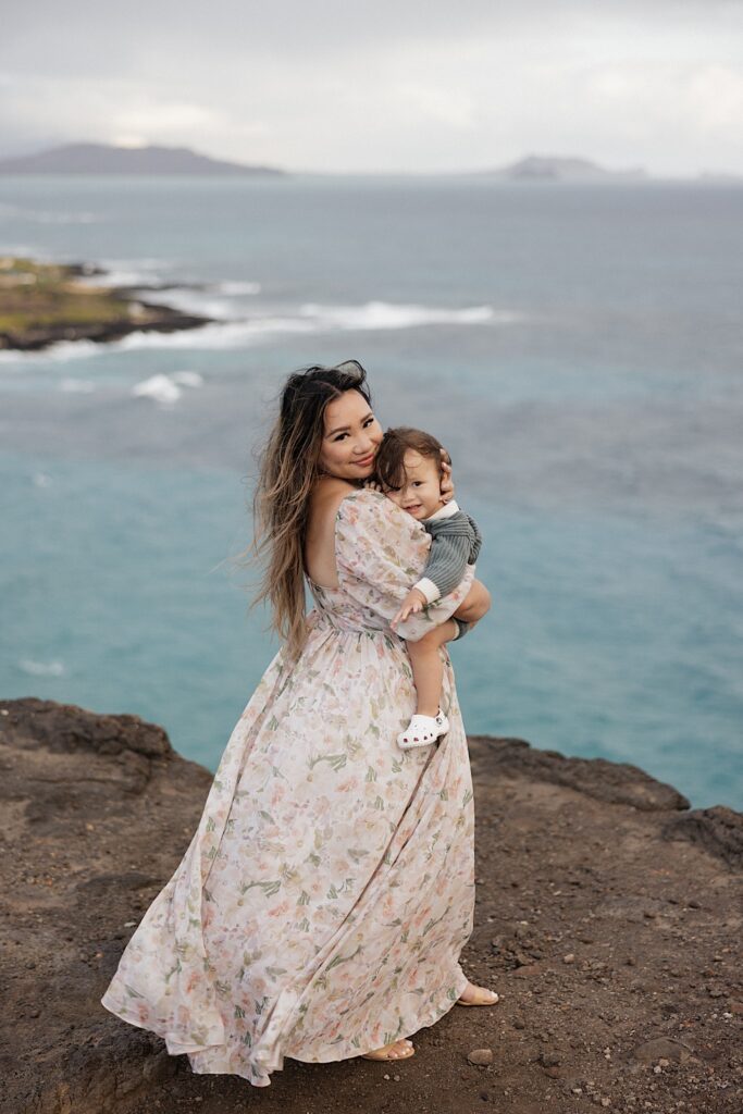 A mother holds a child while standing atop a cliff looking out on the ocean, the mother smiles over her shoulder to the camera as the child also looks at the camera