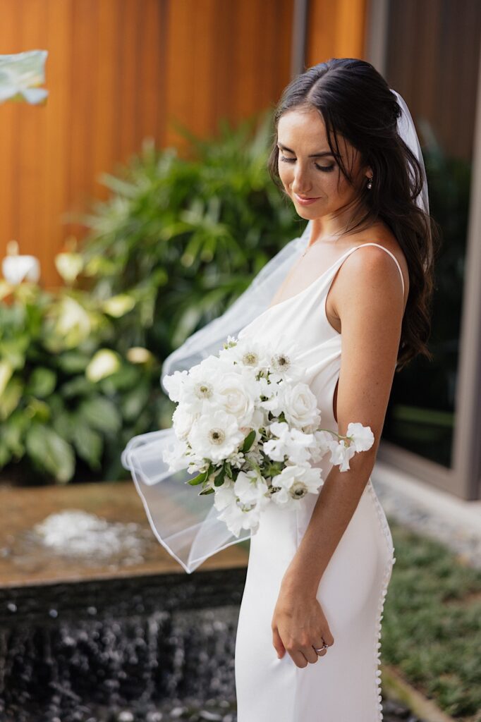 A bride smiles as she looks down towards her bouquet of white flowers that she is holding