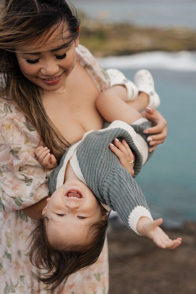 A child smiles at the camera while being held upside down by their mother who is smiling at the child