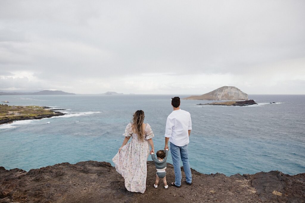 During a family session at Makapuu Lookout on Oahu a mother and father stand atop a cliff and hold their child's hands as they stand between them, all three are looking out over the ocean below them