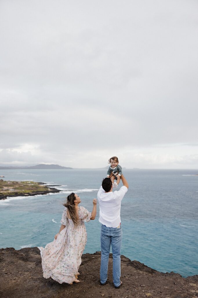 While on a cliff looking out over the ocean a father holds his child in the air while the mother smiles and stands next to them