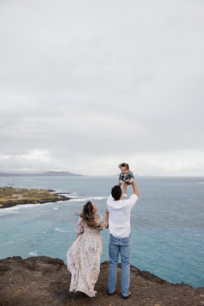 While on a cliff looking out over the ocean a father holds his child in the air while the mother smiles and stands next to them