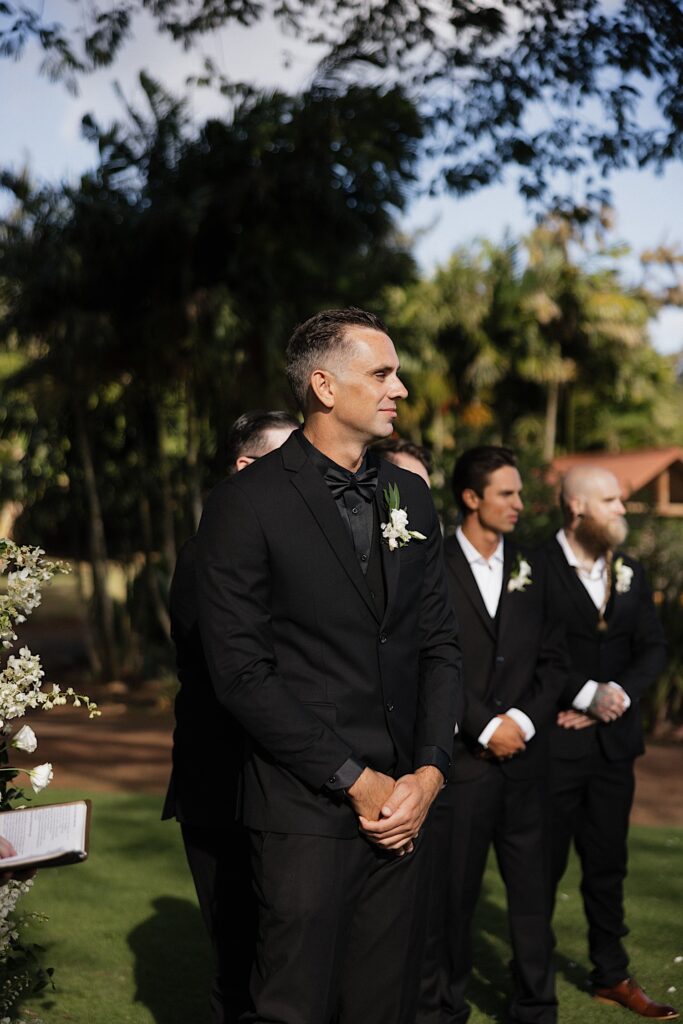 A groom smiles and clasps his hands while looking down the aisle towards the bride who is off camera