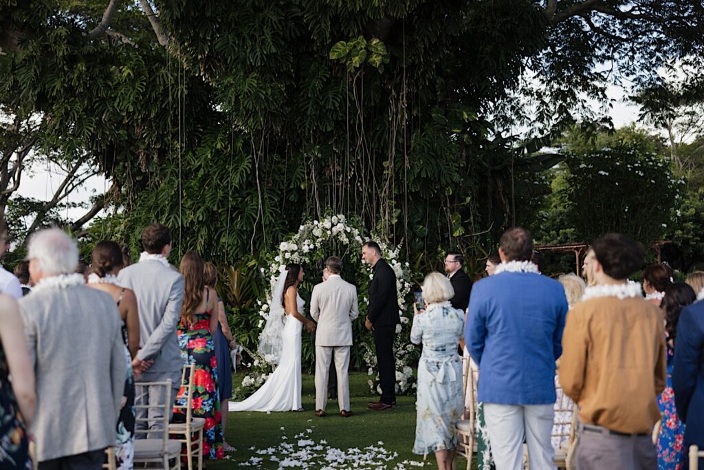 A bride and groom stand during their wedding ceremony at Kukui’ula on Kauai with guests standing and watching as well
