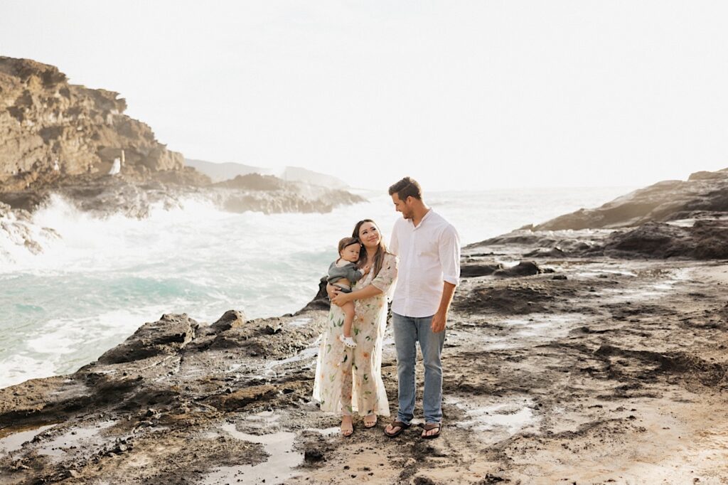 A mother smiles up at her husband while holding their daughter during their family session at Makapuu Lookout on Oahu, next to them are waves crashing on a rocky shore