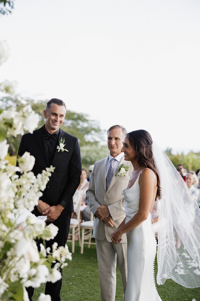 A bride and groom smile during their outdoor wedding ceremony with the bride's father standing and smiling between them
