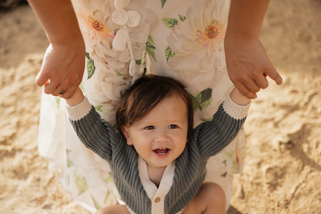 A child looks up at the camera while their arms are held by their mother who is standing above them