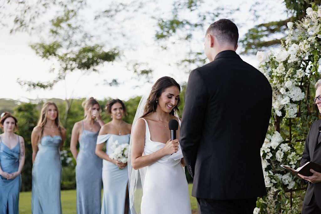 A bride holds a microphone and speaks during her wedding ceremony at the farm at Kukui’ula on Kauai as the groom listens and bridesmaids stand behind 