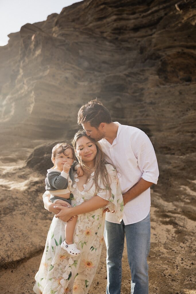 A man kisses a woman on the head as she closes her eyes while holding their child who is looking at the camera, behind them is a large rock formation
