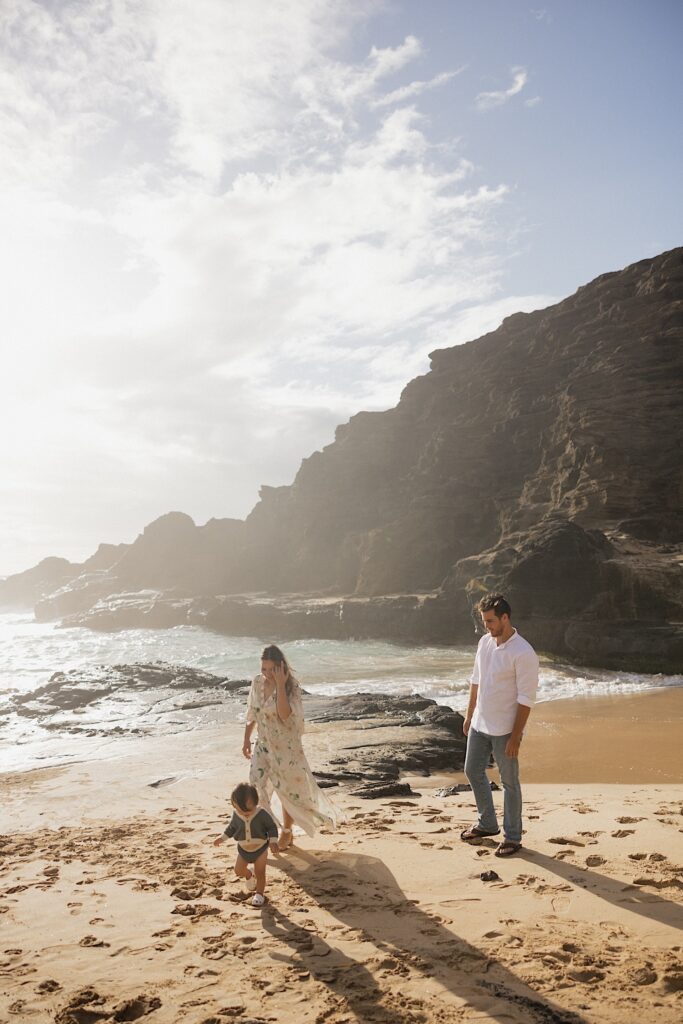 A mother and father walk along a beach following their young child who is wandering around in the sand, behind them is the ocean and a large cliffside