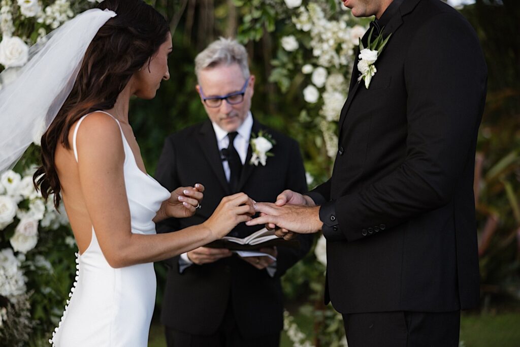 A bride puts a ring on the hand of the groom as their officiant speaks during their wedding ceremony at Kukui’ula on Kauai
