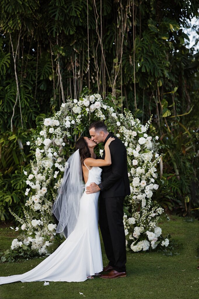 A bride and groom kiss one another in front of a floral archway and large tree