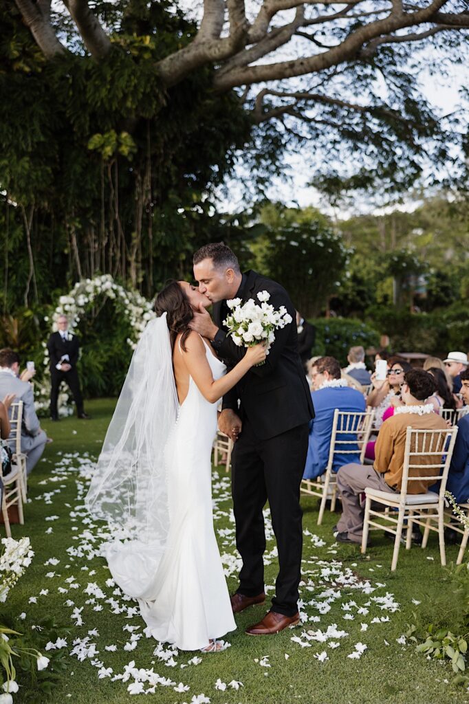 A bride and groom kiss one another while walking down the aisle together after their outdoor wedding ceremony, behind them is a large tree and guests of their wedding