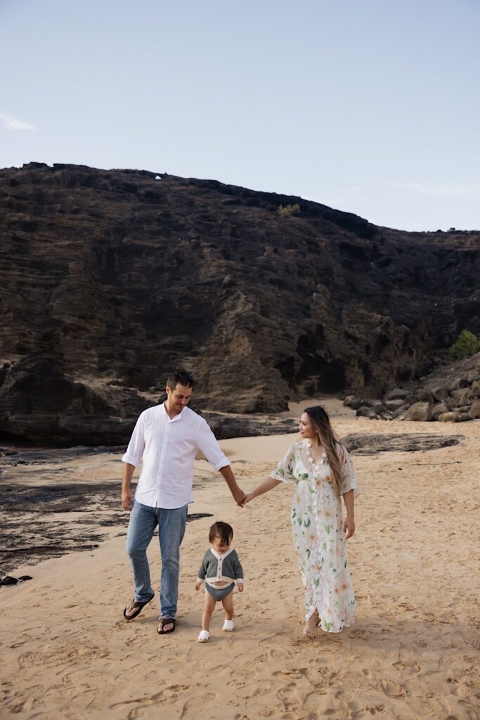 A mother and father walk hand in hand as their young child walks in between them, the three of them are on a beach and there is a large rock formation behind them