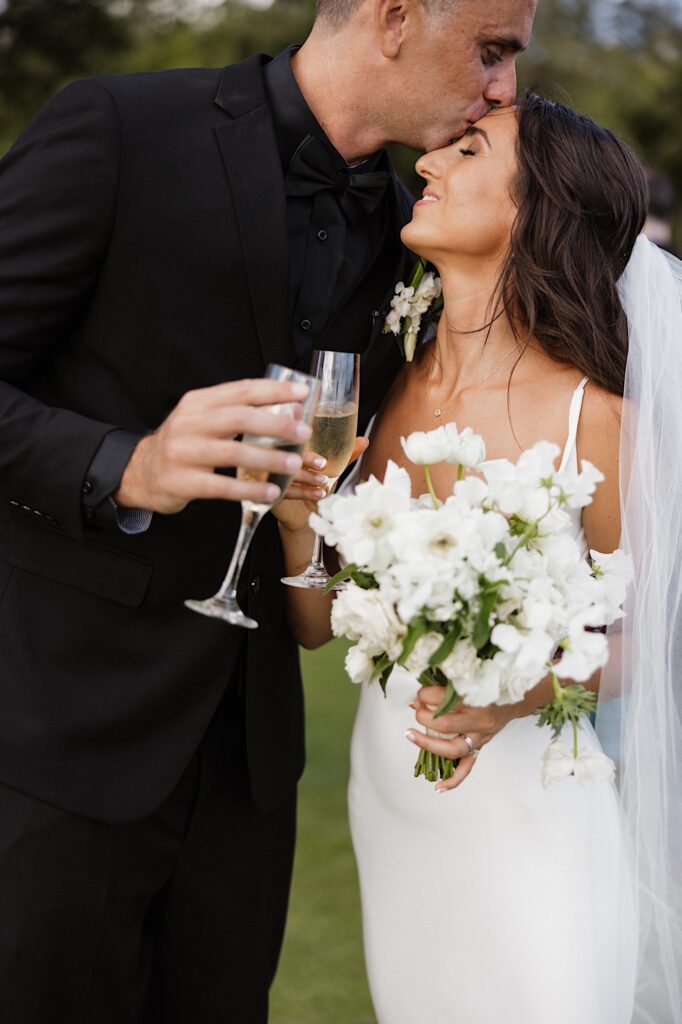 A groom kisses the bride on her forehead as she smiles while they each hold a glass of champagne