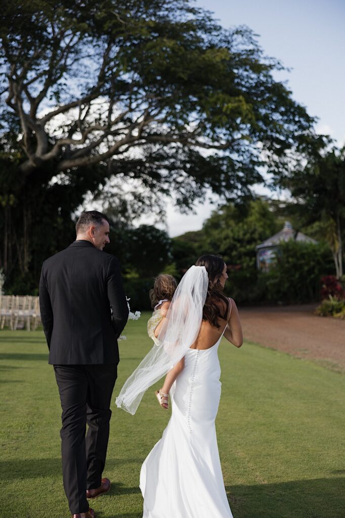A bride and groom walk side by side away from the camera towards a large tree, the bride is carrying a young child in one of her arms