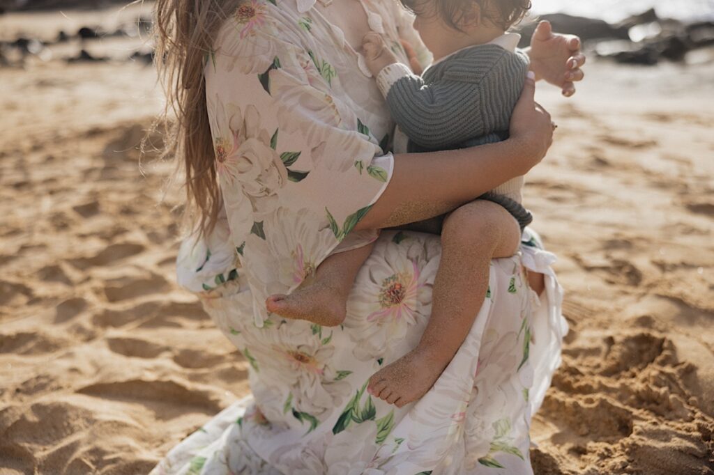 A young child sits on their mother's lap while touching her dress, the mother is sitting on a beach near Makapuu Lookout during their family session on Oahu