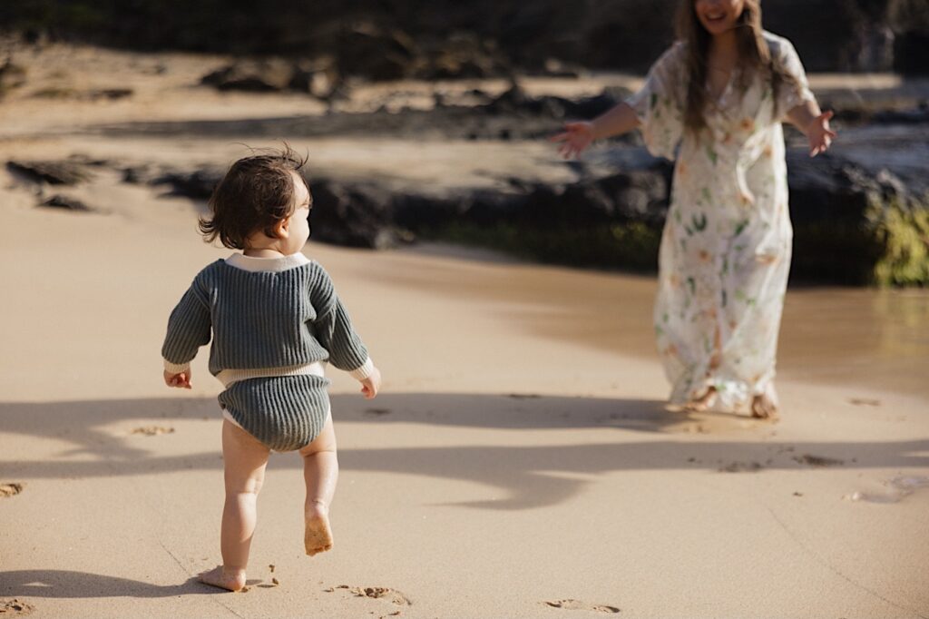 A mother smiles and extends her arms out for her young child to run towards her, they're on a beach during a family session at Makapuu Lookout on Oahu