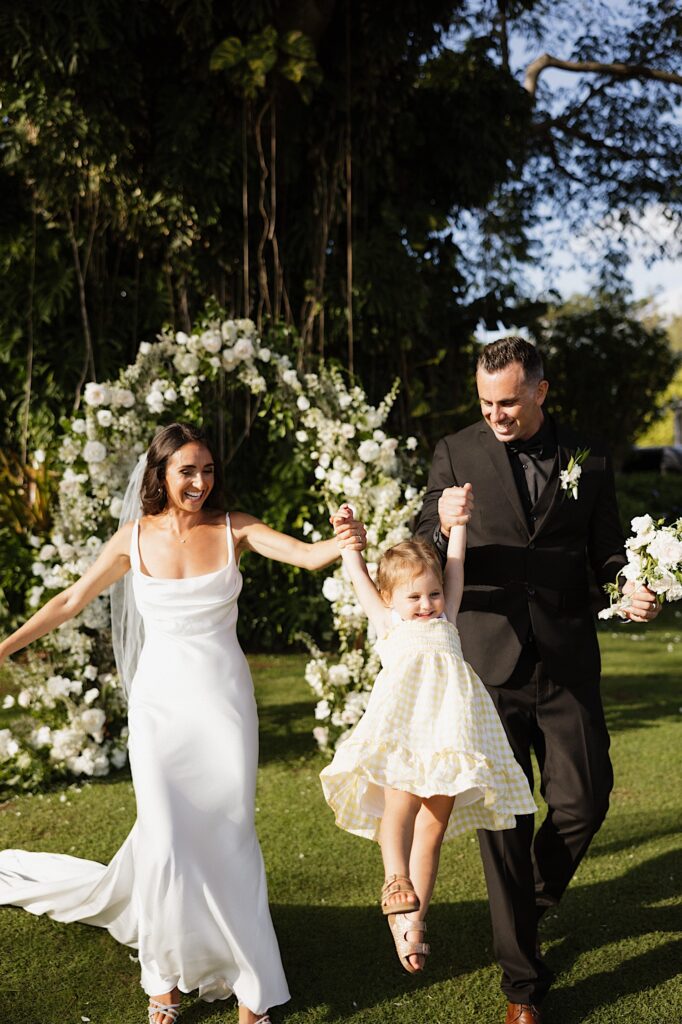 A bride and groom each hold the hand of a young girl and swing her towards the camera as she smiles with a floral archway and massive tree behind them