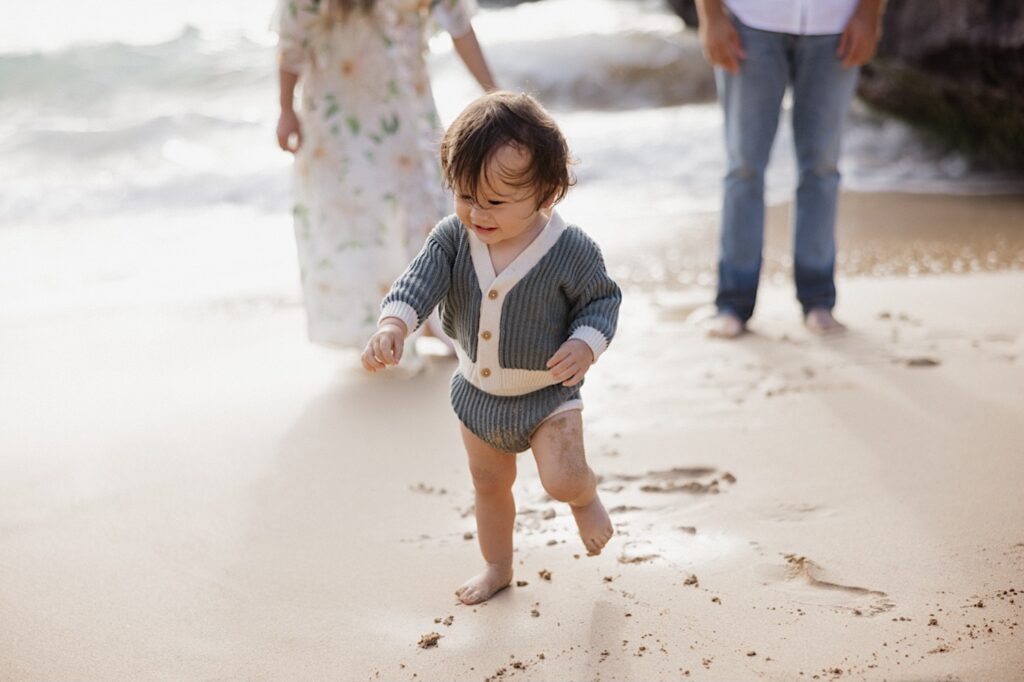 A young child runs in the sand as their parents stand behind them closer to the ocean and watch during their family session at Makapuu Lookout on Oahu