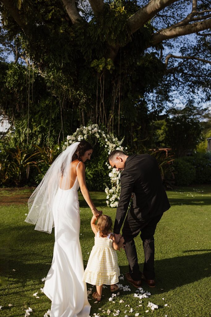 A bride and groom each hold the hand of a young child as they walk together towards their ceremony space 