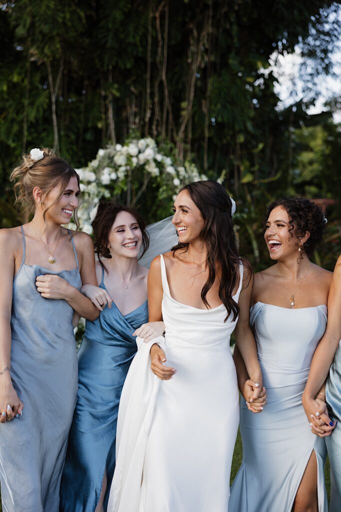 A bride smiles at her bridesmaids who all surround her and smile back while at their outdoor ceremony space