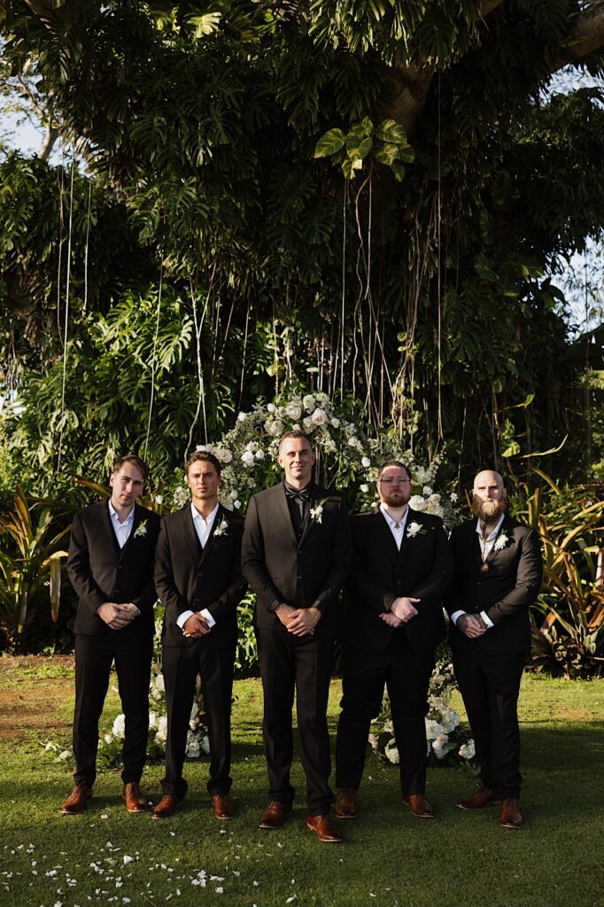A groom poses with his 4 groomsmen who all have their hands clasped and look seriously at the camera, behind them is a floral archway and a large tree