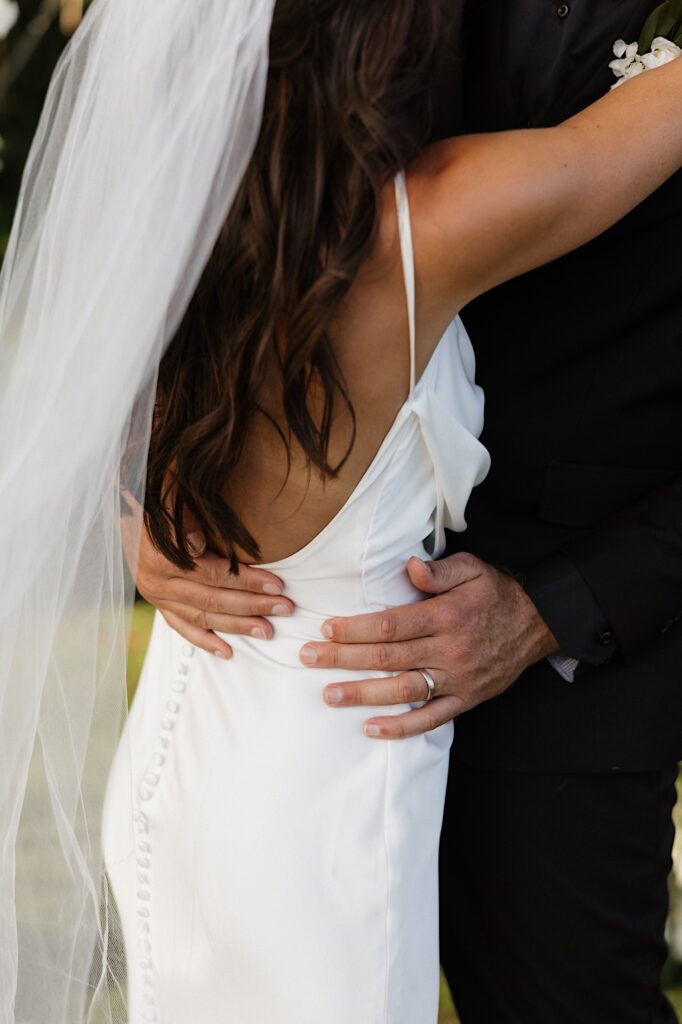 Close up photo of the back of a bride with the groom's hands holding her