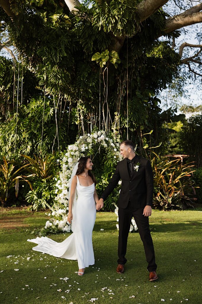 A bride and groom hold hands and walk towards the camera with a floral archway and massive tree behind them