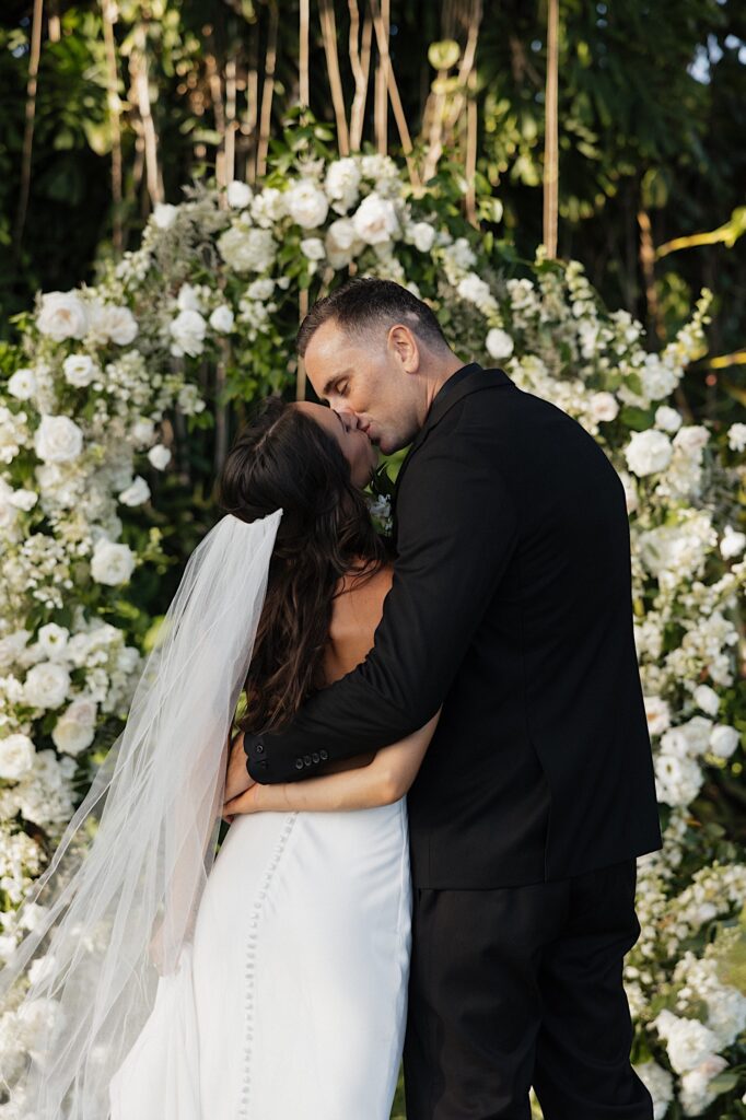 A bride and groom kiss facing away from the camera towards a floral archway and massive tree