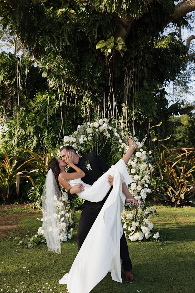 A bride is held in the air and kissed by a groom while standing in front of a floral archway and massive tree