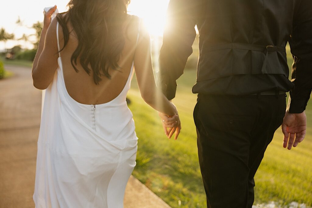 A bride and groom walk hand in hand away from the camera towards the sunset during their wedding day at Kukui’ula on Kauai