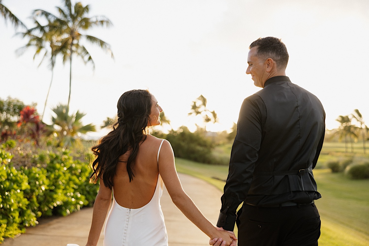 A bride and groom smile at one another while holding hands and walking towards the sunset during their wedding day at Kukui’ula on Kauai