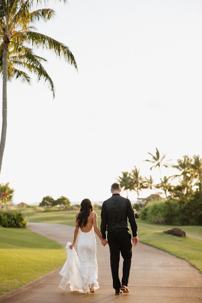 A bride and groom hold hands and walk on a path towards palm trees and the sunset