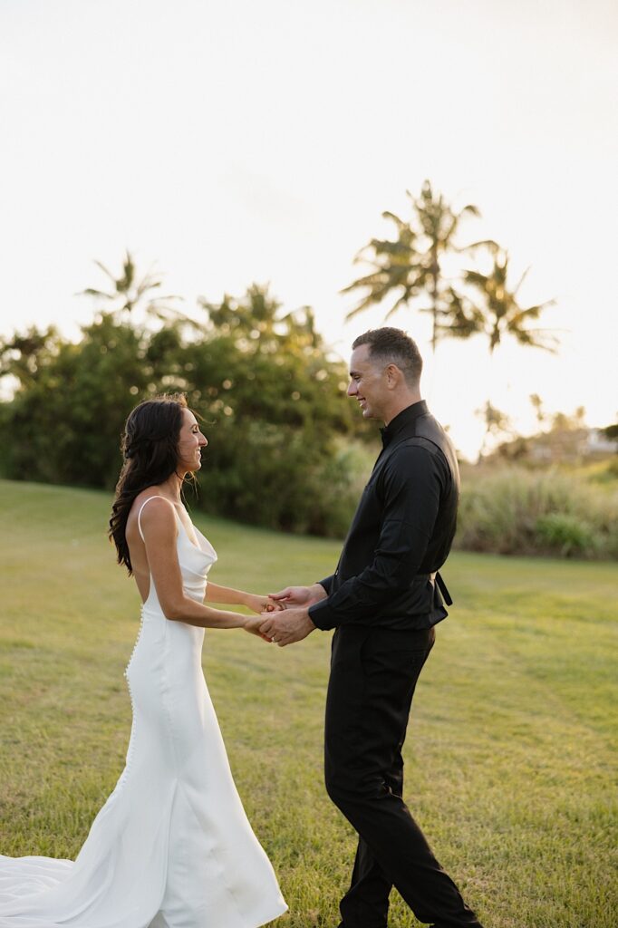 A bride and groom hold hands while dancing in the grass at sunset with palm trees behind them