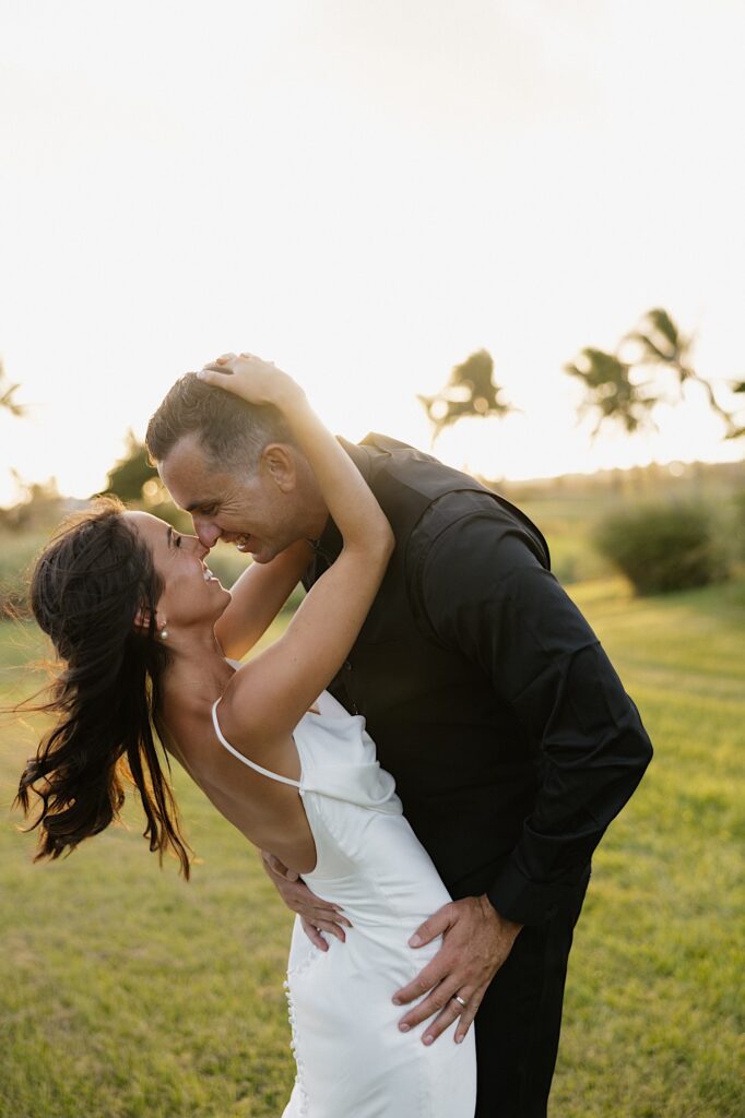 A bride and groom are about to kiss at sunset while standing in a field of grass with palm trees behind them