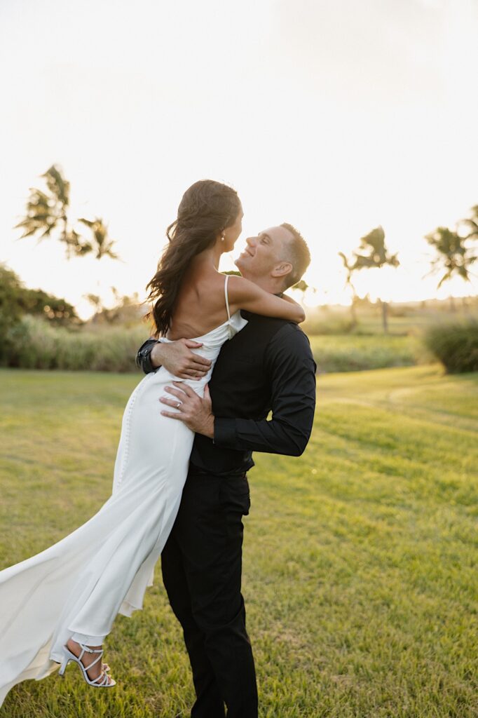 A bride is lifted in the air by the groom who smiles at her while they stand in a field of grass at sunset with palm trees behind them