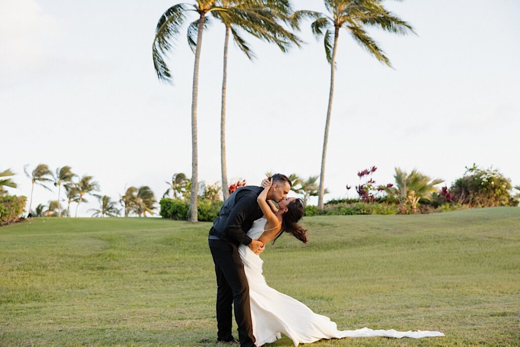 A bride and groom kiss while standing in a grass field as palm trees sway behind them during their wedding day at Kukui’ula on Kauai