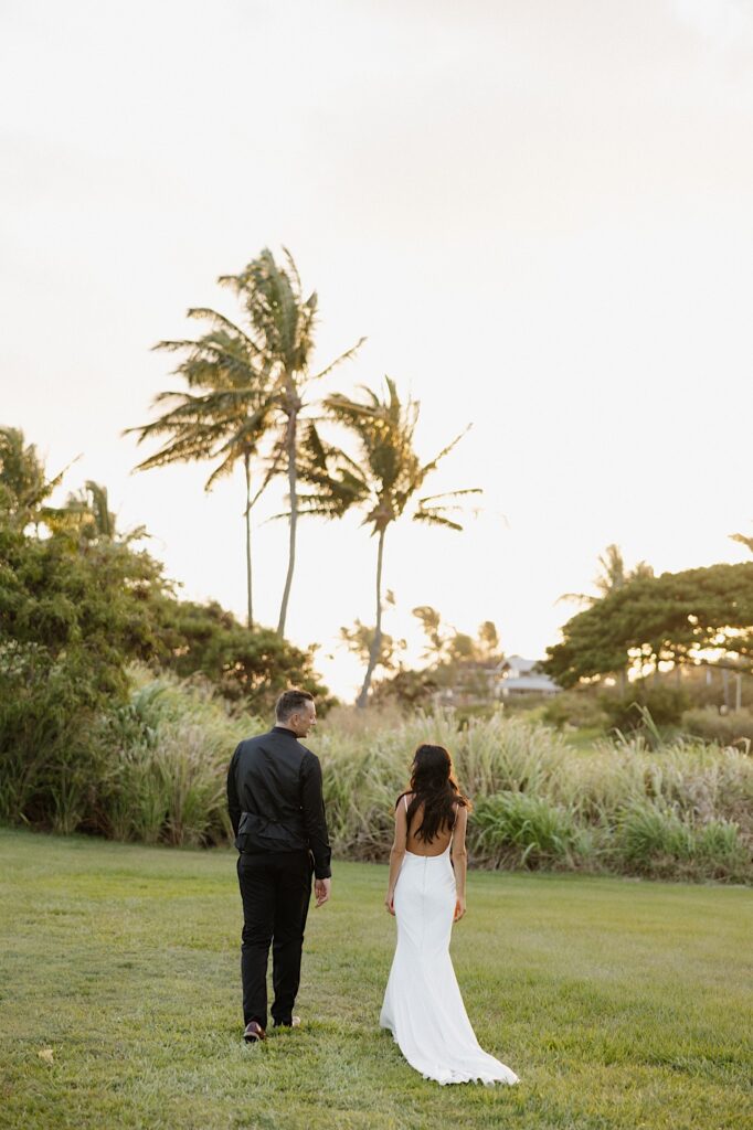 A bride and groom walk side by side in a grass field towards palm trees away from the camera